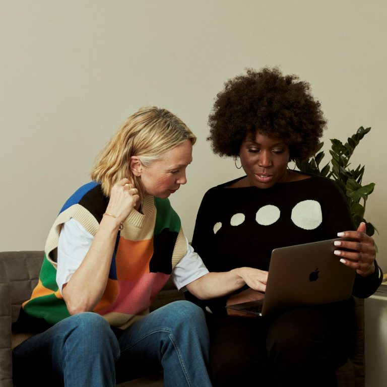 Two women sitting in a modern office lounge having a conversation, one holding a notebook and the other holding a coffee mug.