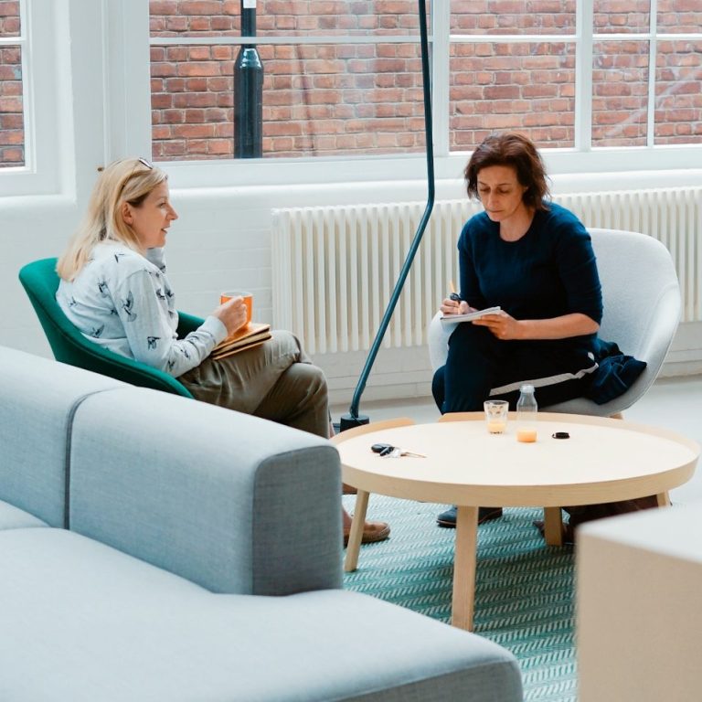 Two women sitting in a modern office lounge having a conversation, one holding a notebook and the other holding a coffee mug.