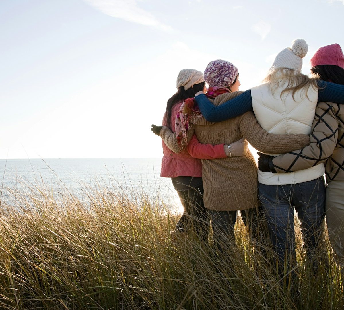 Four people standing arm-in-arm, facing the ocean on a sunny day, dressed in winter clothing and hats, surrounded by tall beach grass.